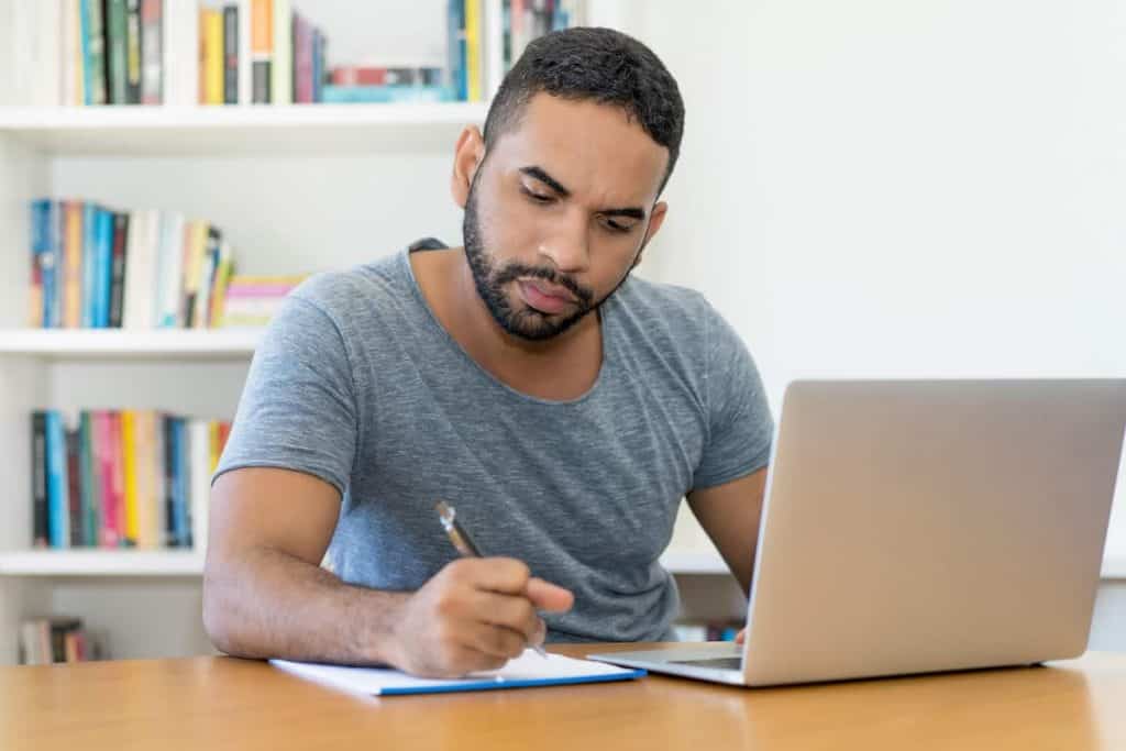 man working on a laptop computer and writing notes on a pad of paper